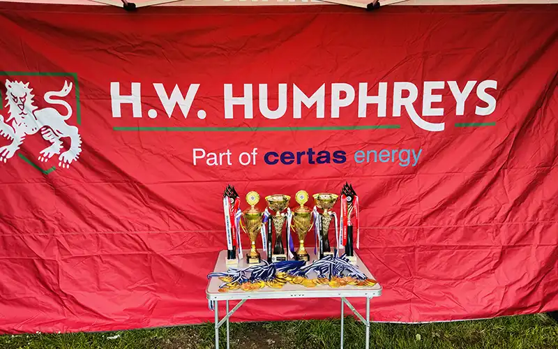 A red banner with HW Humphreys, part of Certas Energy text in front of a table of sports trophies and medals.
